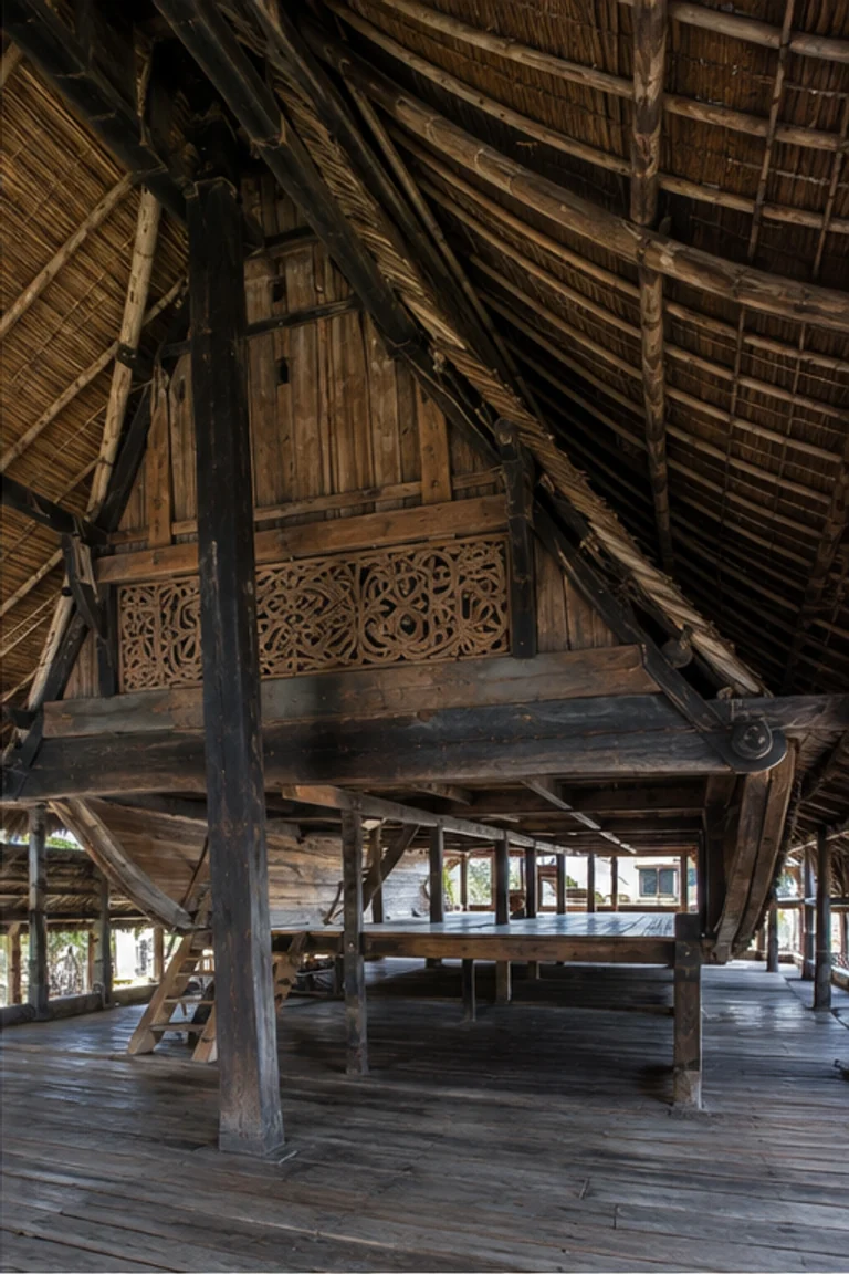 People-free photorealistic vertical Batak Toba ruma with massive raised hardwood frame, boat-like section, steep saddle roof, dark undivided family floor, hearth, woven mats, chests, baskets, and restrained red-black-white gorga carving lit from the gable.