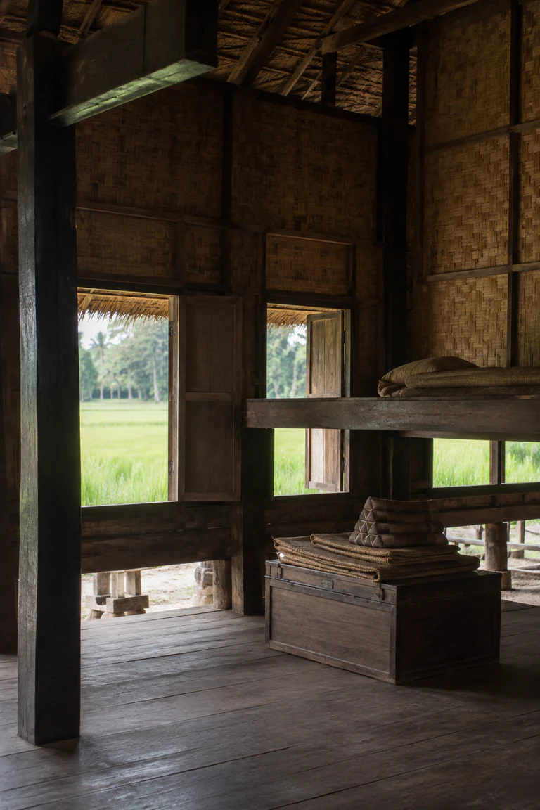 Photorealistic vertical view inside a traditional Khmer raised house with dark timber posts, woven bamboo wall panels, reed mats, a low chest, open shutters, and humid green fields beyond the shaded undercroft.