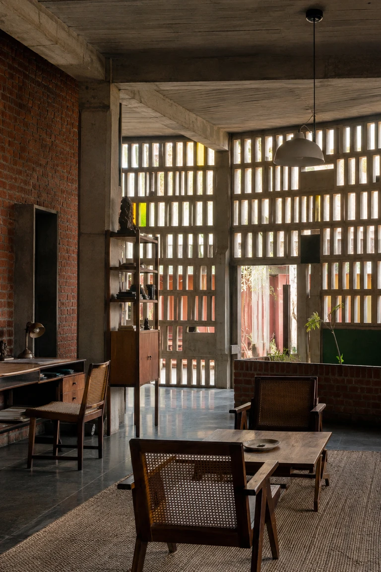 Chandigarh modernist room with board-marked concrete, exposed brick, deep perforated screens, timber-and-cane chairs, modular desk, woven rug, and filtered Indian daylight.