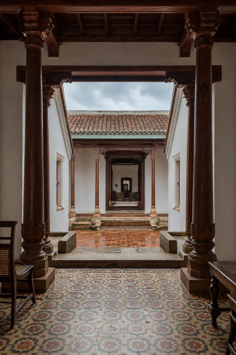 Photorealistic vertical axial view through a Chettinad house thinnai and successive courts, with carved Burmese-teak columns, patterned Athangudi tile, lime-plaster walls, stone thresholds, restrained timber furniture, and a bright square of monsoon sky.