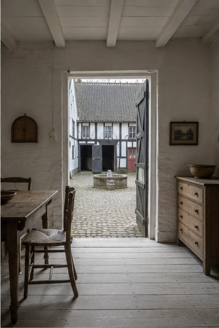 Photorealistic vertical Danish four-wing farm viewed from a compact limewashed family room through a timber doorway into the protected rectangular yard, with half-timbered barn and stable wings, scrubbed table, painted cupboard, ceramics, and cool northern daylight.