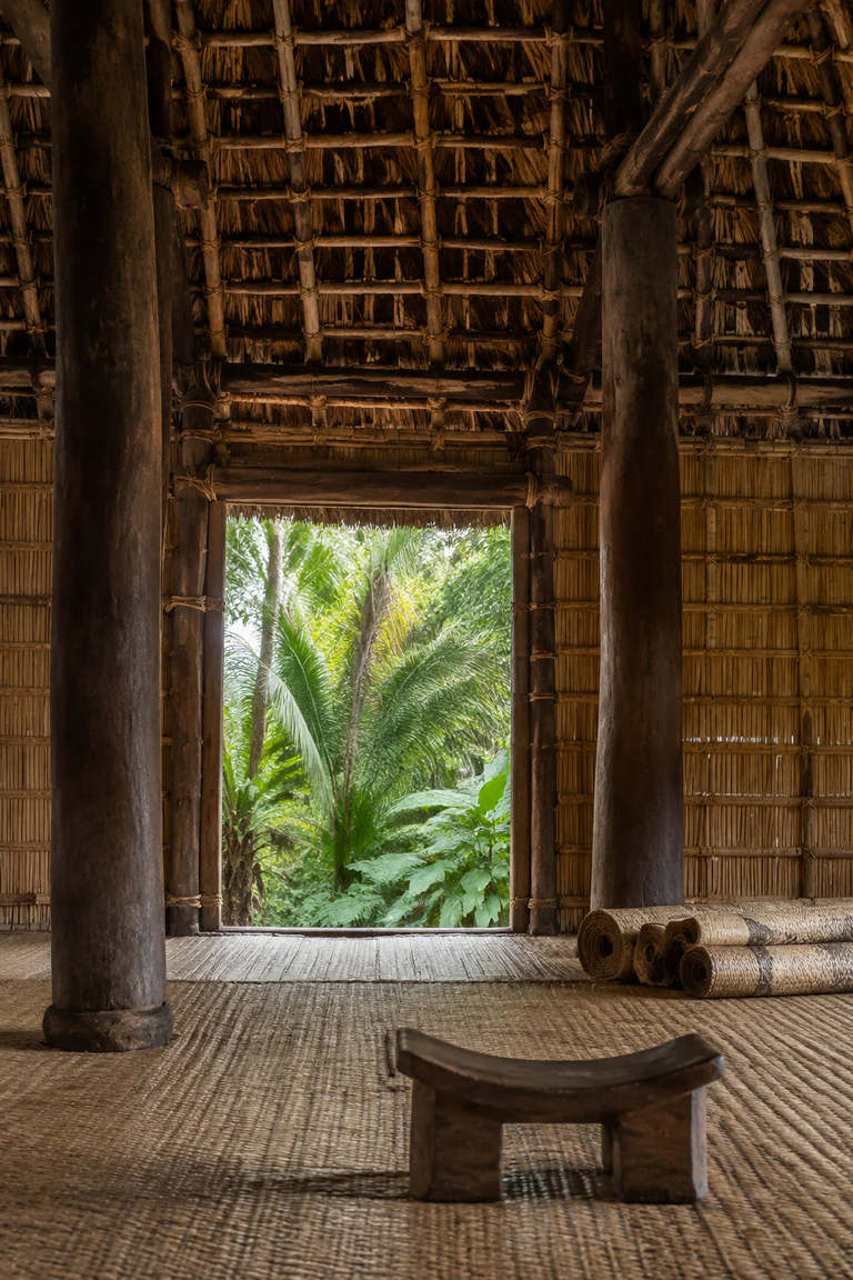 Photorealistic vertical Fijian bure with massive hardwood posts, layered reed walls, high sennit-lashed thatch, pandanus mats, barkcloth rolls, low timber headrests, and a broad doorway framing humid tropical vegetation.