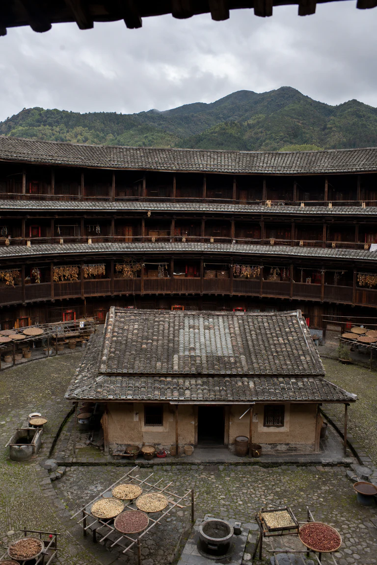 Photorealistic vertical interior courtyard of a circular Fujian tulou with a massive rammed-earth ring, three levels of timber galleries, equal household bays, clay-tiled eaves, domestic drying racks, and a central ancestral hall.