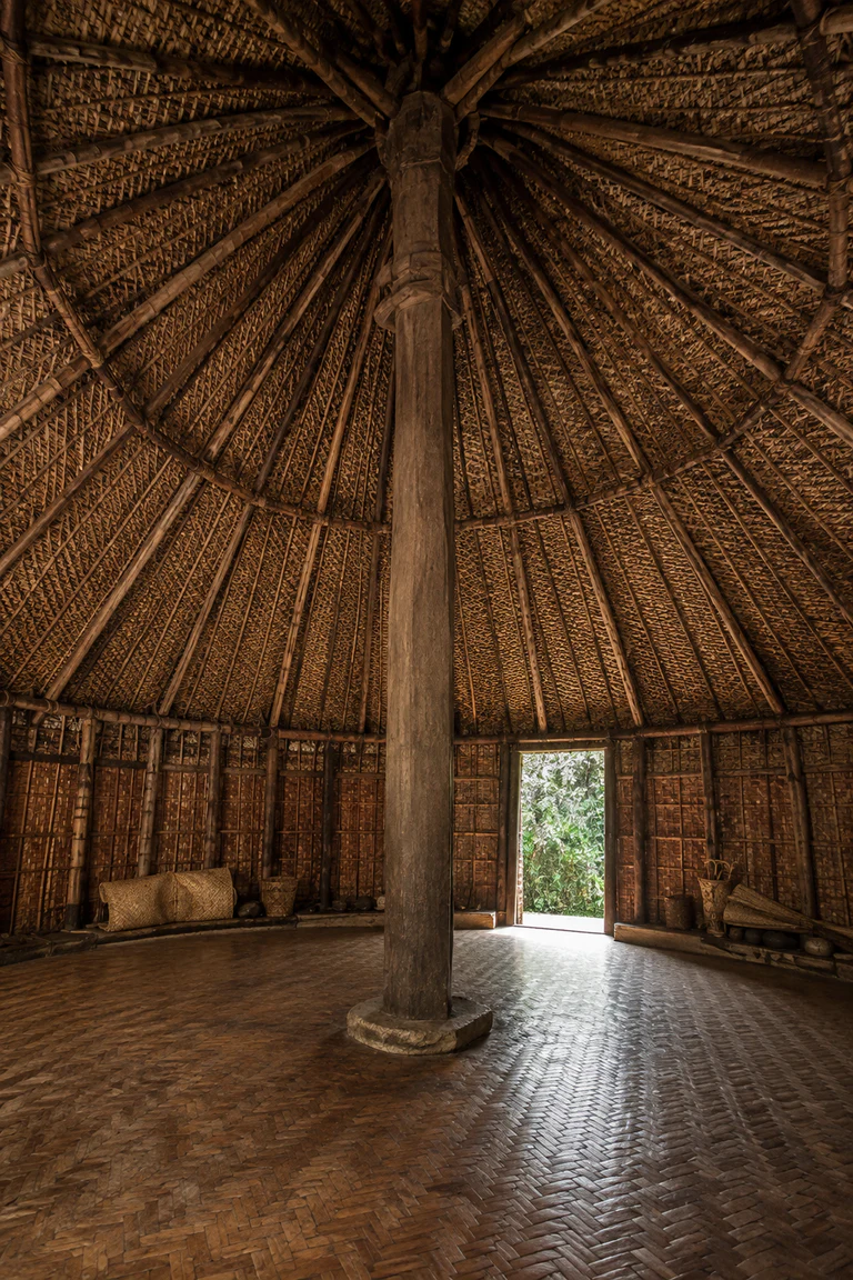 Photorealistic vertical Kanak grande case interior with a soaring central hardwood post, radiating roof members, woven pandanus mats, barkcloth bundles, low perimeter objects, tall conical thatch, and one filtered doorway to tropical vegetation.