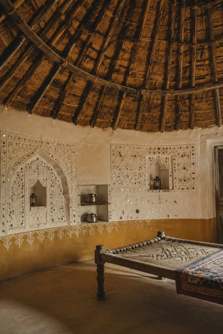 Photorealistic vertical Kutch bhunga interior with sculpted lipan kaam relief and small inset mirrors, white and ochre earthen walls, conical timber-and-thatch roof, built-in niches, embroidered textiles, low charpoy, and warm desert light.