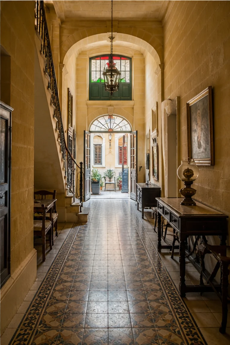 Photorealistic vertical Maltese townhouse with a long limestone entrance hall, patterned cement tile, steep stair, formal room opening, coloured light from an enclosed painted-timber gallarija, dark cabinets, brass, lace, and a bright rear court.
