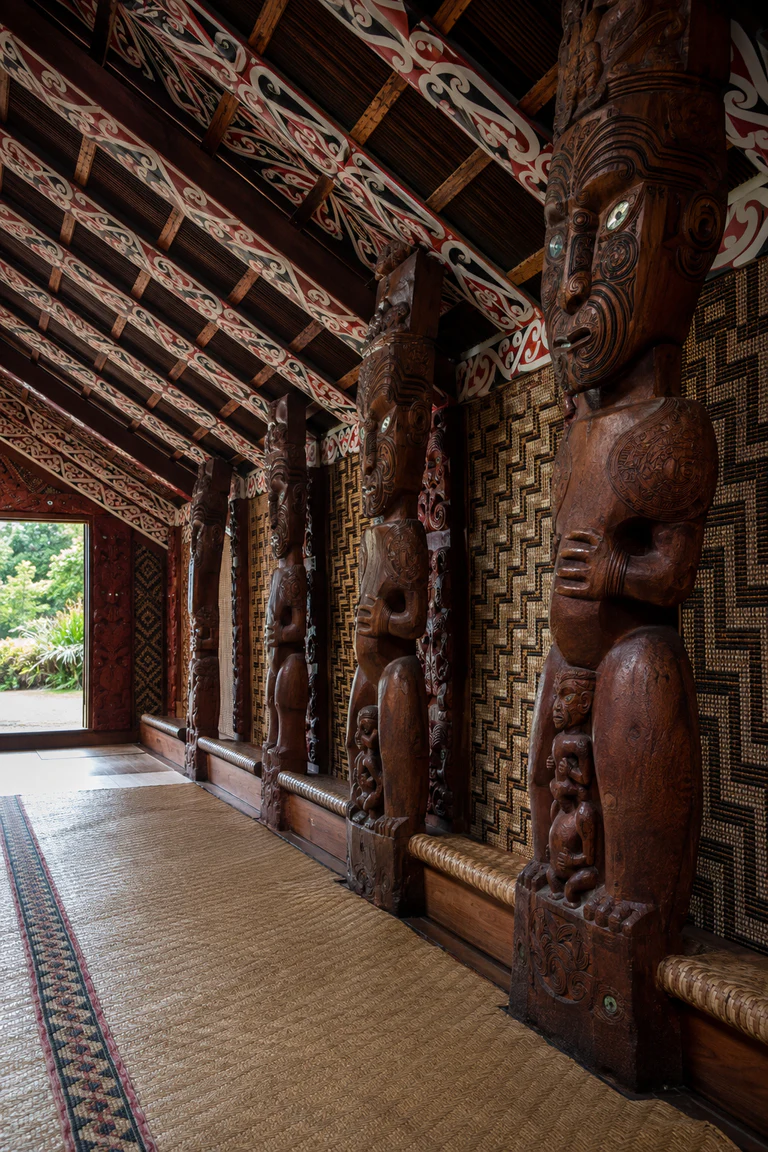 Photorealistic vertical Māori wharenui with aligned carved poupou ancestor figures, red, black, and white kōwhaiwhai rafters, woven tukutuku panels, central aisle, flax mats, low perimeter seating, and daylight from the open porch.