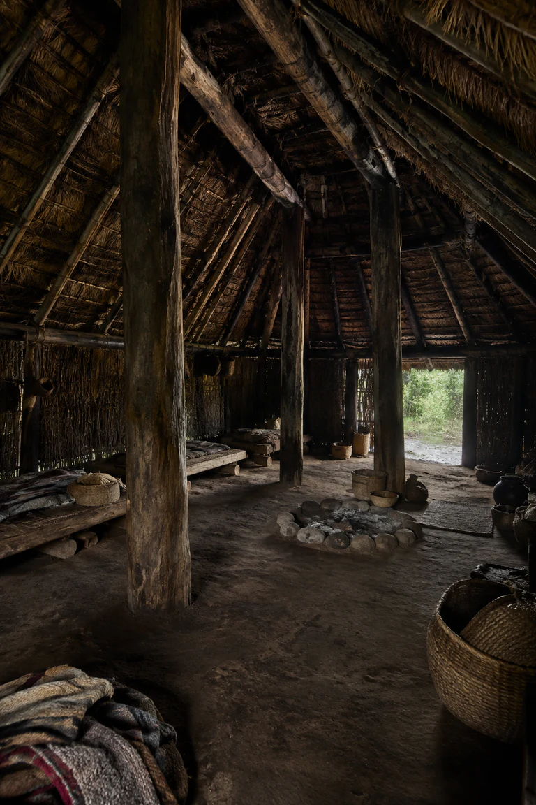 Photorealistic vertical Mapuche ruka with heavy timber posts, dense coirón-thatch roof, a central hearth, packed-earth floor, low sleeping platforms, woven wool, baskets, food-storage vessels, and rain-softened daylight at a broad entrance.
