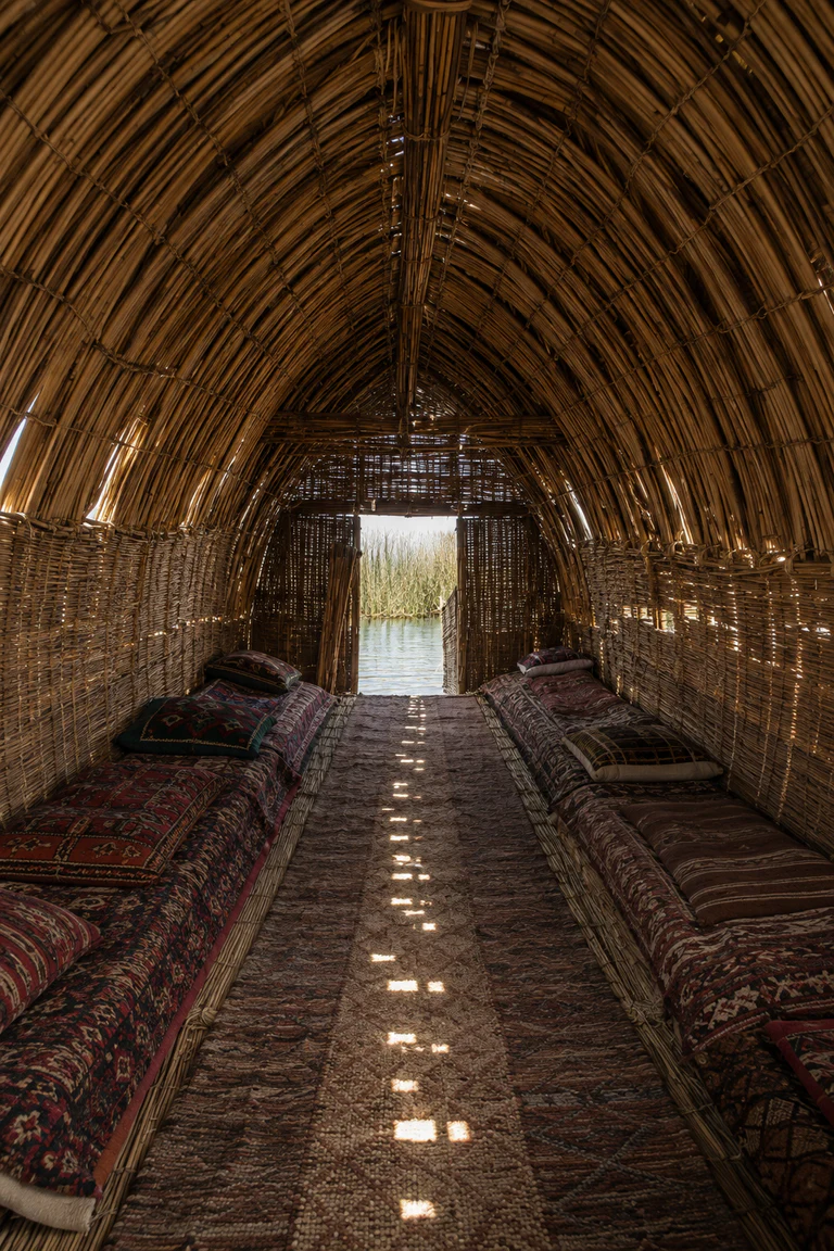 Photorealistic vertical interior of a southern Iraqi mudhif with repeated bundled-reed arches, woven mat walls, low mattresses and rugs, patterned slivers of daylight, and the Mesopotamian marsh visible through the entrance.