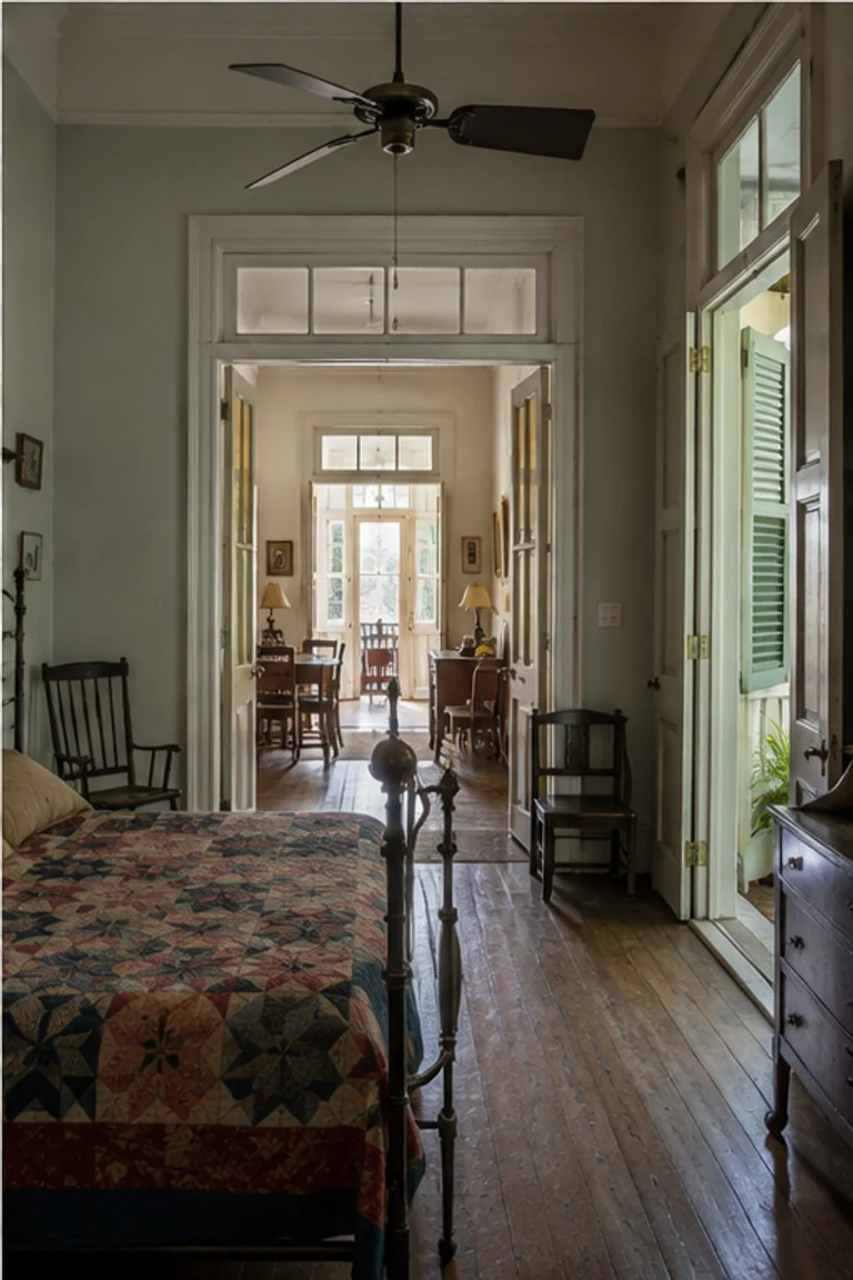 Photorealistic vertical New Orleans shotgun house with aligned doors through a narrow hall-free sequence of front room, bedroom, and rear kitchen, high ceilings, transoms, pine floors, painted trim, iron bed, patterned quilt, family photographs, and humid porch light.
