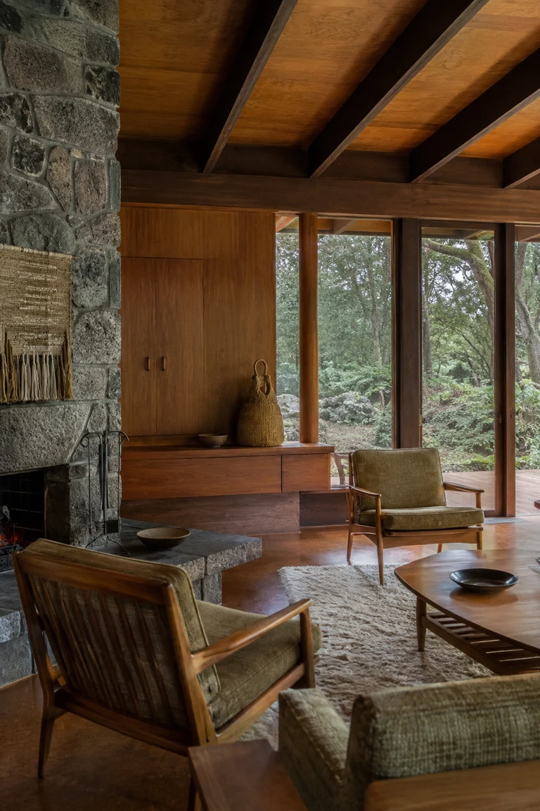 New Zealand mid-century room with exposed timber rafters, rimu cabinetry, volcanic stone hearth, wool lounge chairs, flax weaving, and broad native-bush windows.