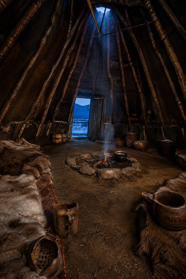 Photorealistic vertical turf-covered Sámi goahti with a curved birch frame, central stone hearth and smoke opening, reindeer hides, low bedding, carved wooden vessels, packed-earth floor, and blue Arctic twilight at the doorway.