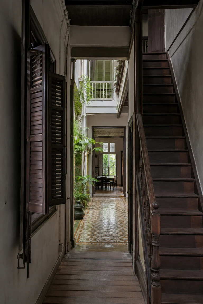 Early twentieth-century Singapore shophouse interior with a long narrow axis, timber shutters and boards, rain-wet patterned tile in a central airwell, steep timber stair, dark furniture, and a rear domestic room beyond.