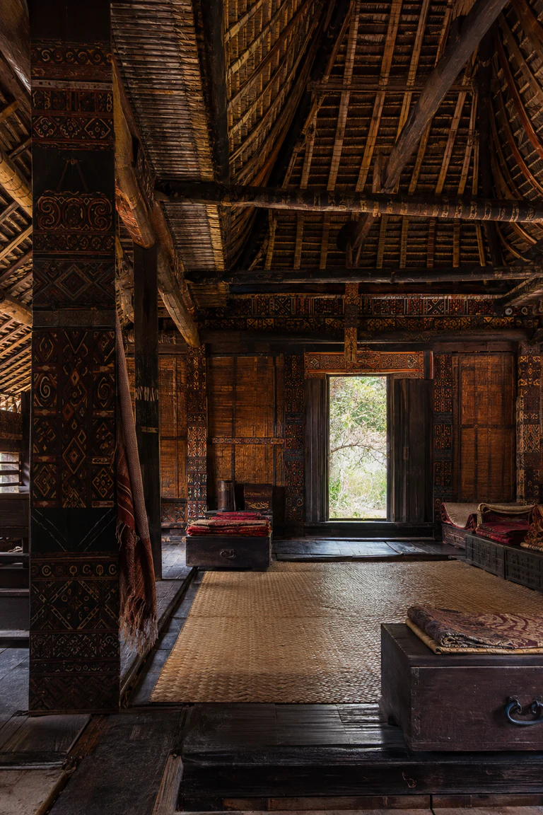 Photorealistic vertical Toraja tongkonan interior with a dark raised chamber beneath a curved bamboo roof, red, black, and yellow carved structural panels, a central mat zone, storage chests, woven cloth, and cool mountain daylight.
