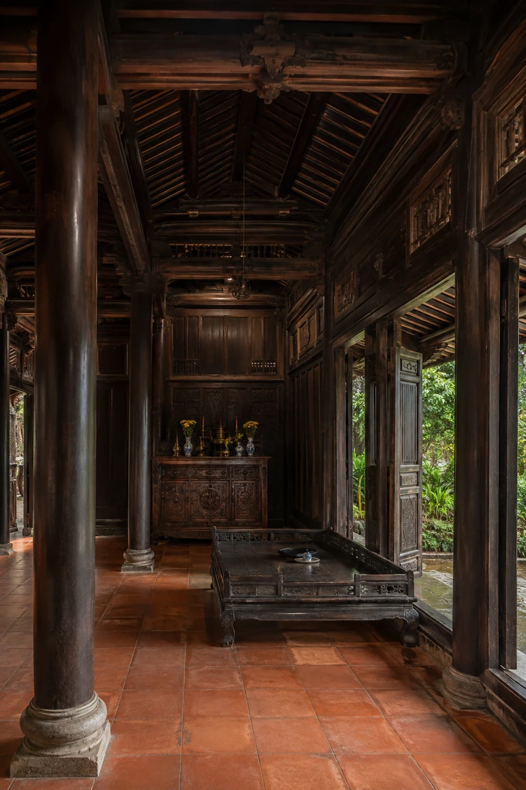 Traditional Huế rường house interior with polished dark timber columns, exposed joinery, central carved altar cabinet, low daybed, terracotta floor, and open doors framing a rain-green garden.